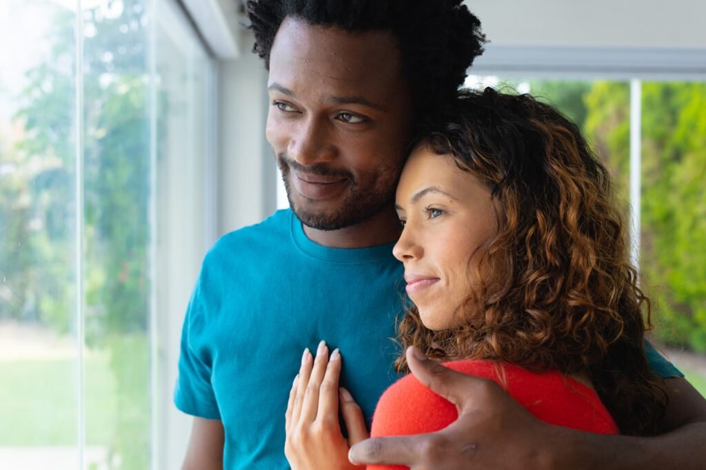 Romantic multiracial couple embracing while looking out of window together at home